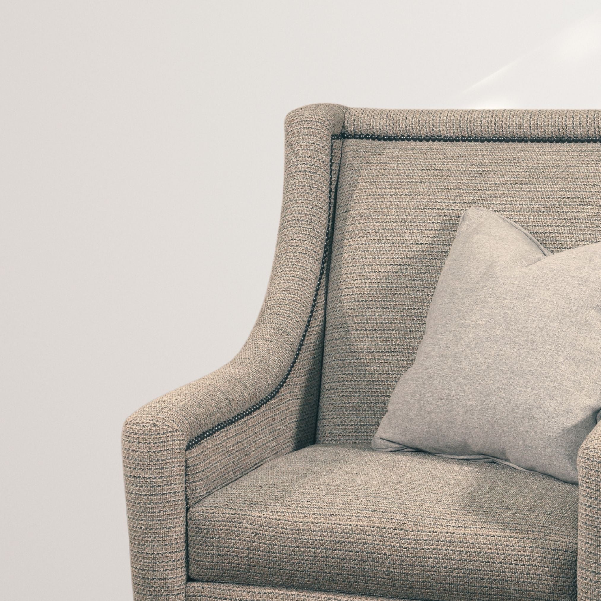 A close-up of a modern, light gray upholstered armchair with subtle texture and a sleek, curved silhouette. The chair features dark piping along the edges, adding a refined touch. A matching throw pillow rests on the seat, enhancing its comfort and style.