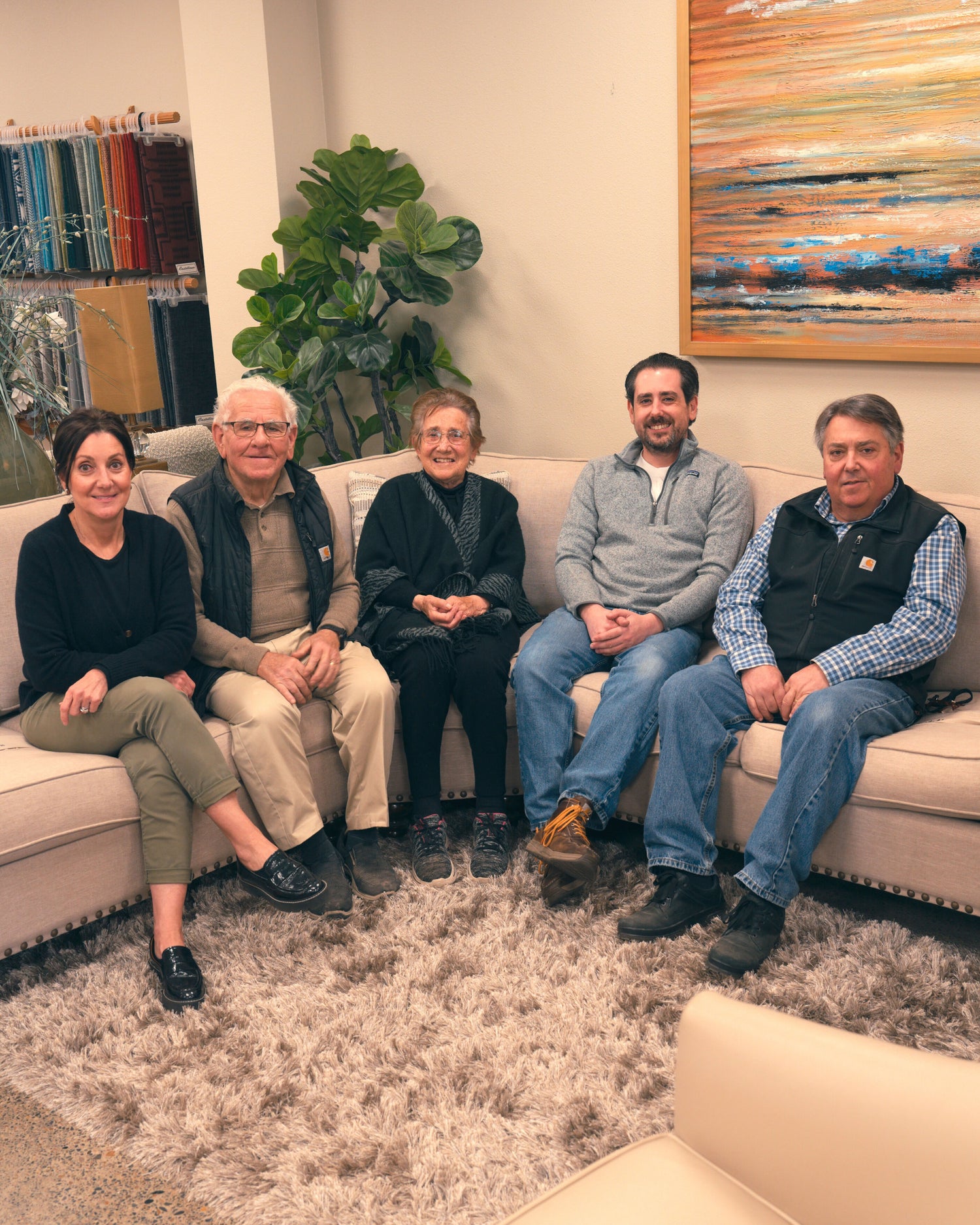 The Castellano family, representing three generations of craftsmanship, sits together on a light-colored sofa in a warm, welcoming space. Behind them, vibrant textiles and an abstract painting add to the cozy ambiance. The family looks proud and connected, reflecting the legacy of Castellano Custom Furniture, a business built on tradition, collaboration, and personalized design.