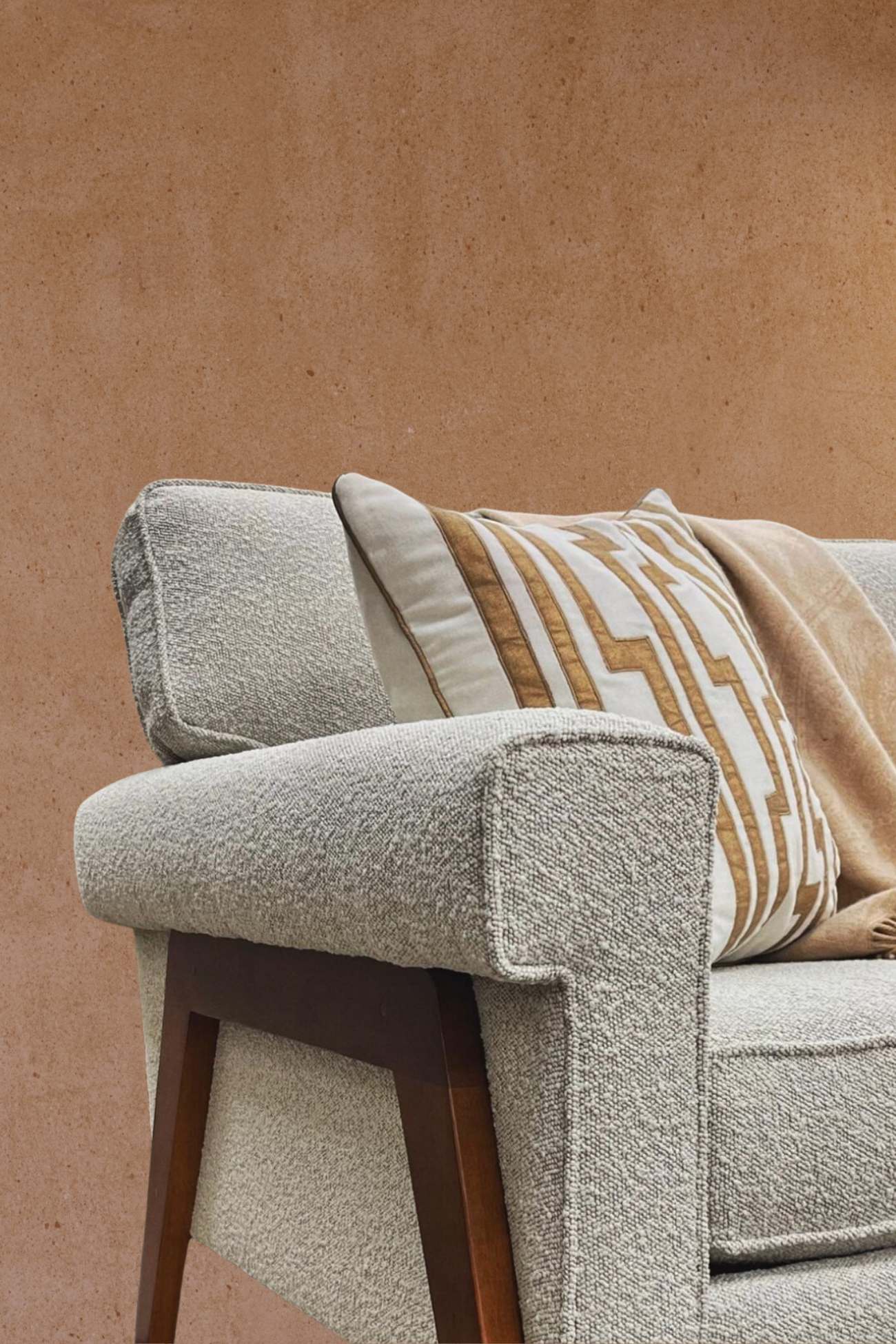 A modern light gray armchair with textured upholstery and sleek wooden legs. A patterned throw pillow in earthy tones and a cozy beige blanket drape across the seat, adding warmth and style to the minimalistic design.