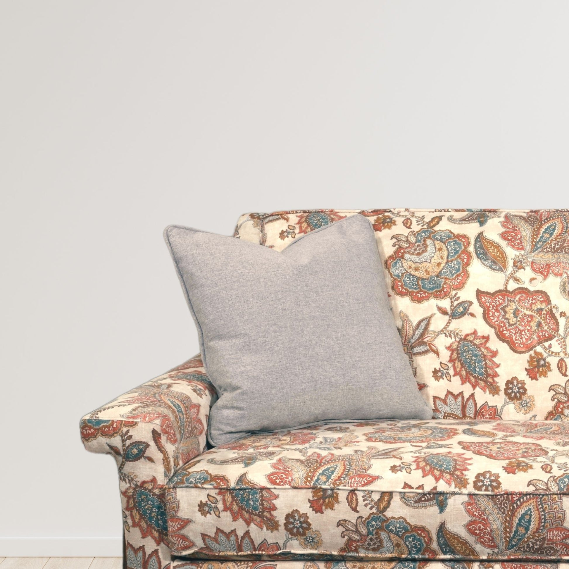 A vintage-inspired loveseat featuring a colorful paisley pattern in shades of blue, red, and brown on a cream background. A soft, light gray throw pillow sits on the seat, adding a touch of modern contrast to the classic, detailed upholstery.